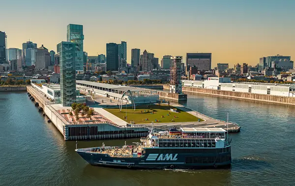 A blue cruise ship with the "AML" logo sails near a modern quay with green spaces, surrounded by the skyscrapers of a city under a clear and sunny sky.
