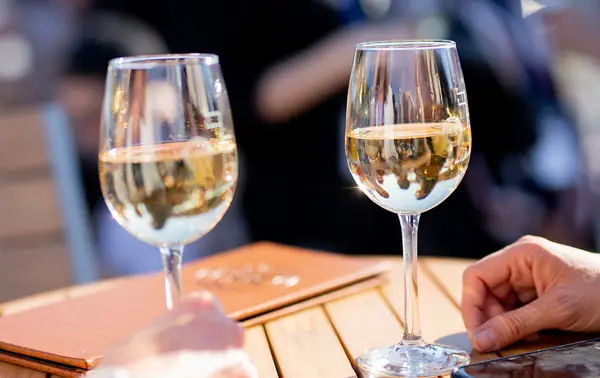 Two glasses of white wine placed on a wooden table, with leather menus and a hand visible in the foreground. The background is blurred, suggesting a bright and sociable outdoor atmosphere.