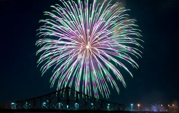 A massive multicolored firework bursts in the night sky above an illuminated bridge, creating a spectacular and vibrant scene.