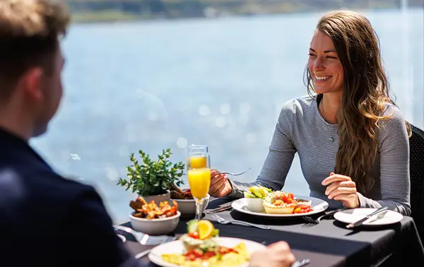 Two people sharing a meal at an elegant table with a view of the water. A woman smiles while holding a fork, with colorful dishes, a glass of mimosa, and table decorations.
