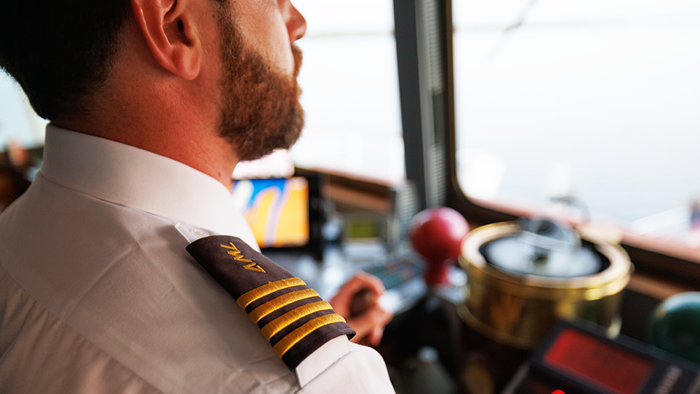 A ship captain in uniform, seen from behind, overlooking the navigation equipment and the St. Lawrence River from the deck of a ship.