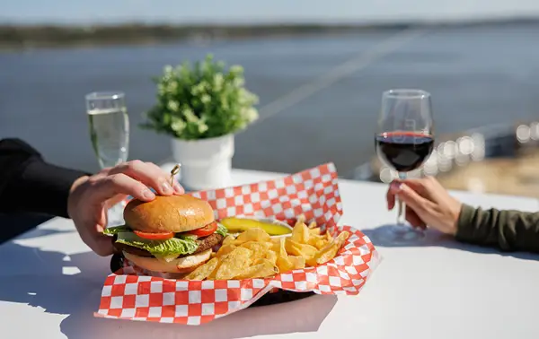 A plate with a hamburger, chips, and a pickle on a white table with a red-and-white checkered pattern. One person holds a glass of red wine, another touches the burger. A river and a potted plant are visible in the background.