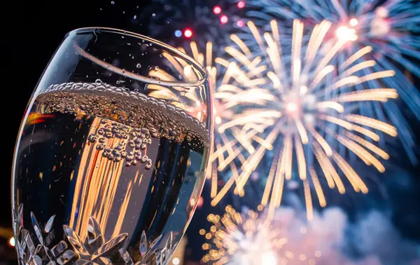 A close-up of a sparkling champagne flute in the foreground, with vivid fireworks lighting up the night sky in the background.