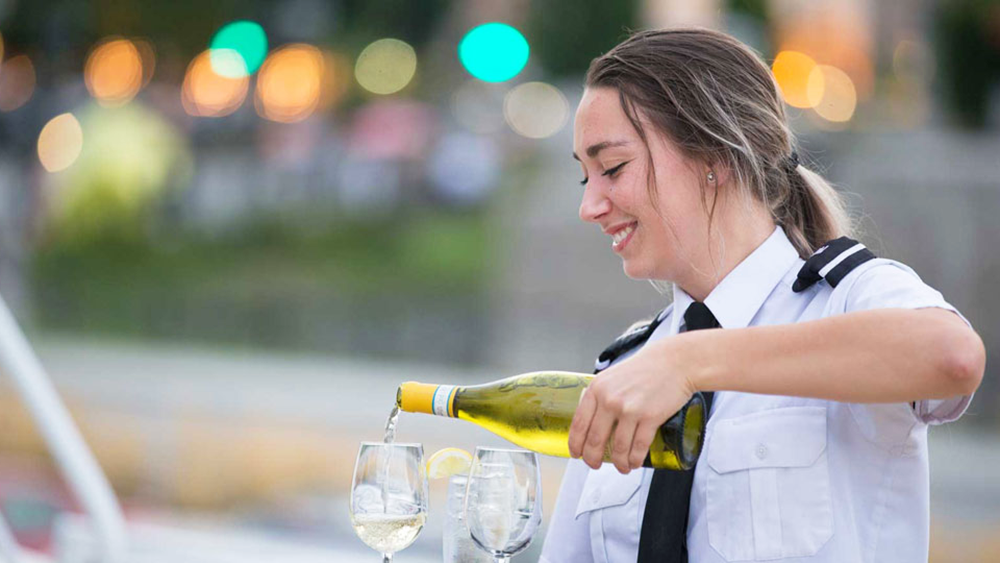 A smiling waitress pouring white wine into two glasses on a table.