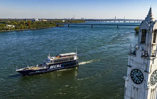 Aerial view of a cruise ship sailing on a river, with a large white clock tower in the foreground and a bridge in the background under a clear sky.