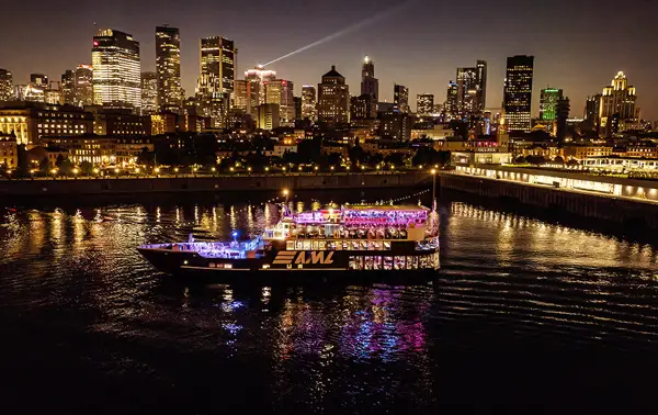A stunning nighttime view of an illuminated cruise ship sailing on a river, with a sparkling city skyline in the background featuring skyscrapers and urban lights.