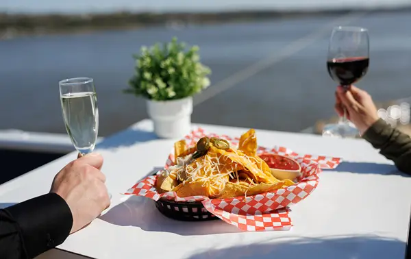A white table with a basket of nachos topped with cheese and jalapeños, served with red sauce. One person holds a glass of red wine, and another a glass of white wine, with a river and a plant in the background.