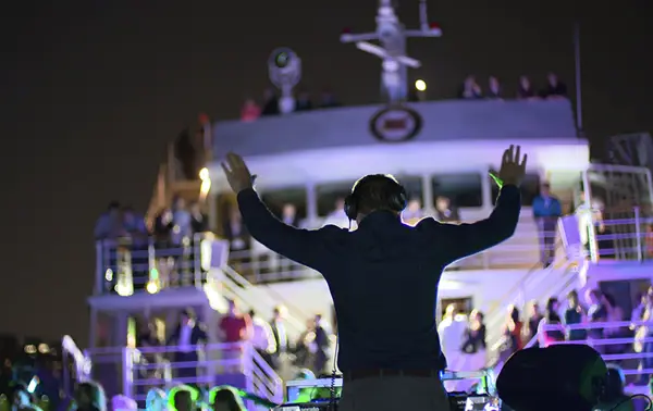 A DJ hosts a lively party on the deck of a boat, arms raised, with a crowd of attendees and colorful lights illuminating the scene at night.