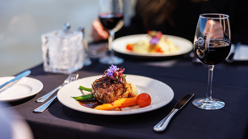 Un plat de steak gourmet garni d'une fleur, accompagné d'un verre de vin rouge, avec vue sur le cadre serein d'une vue sur l'eau.