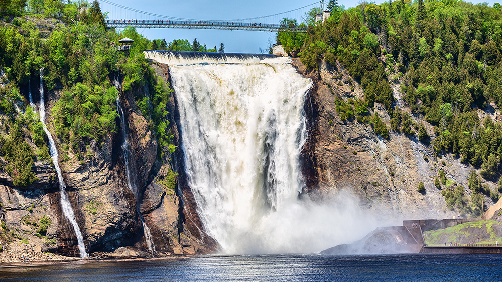 Montmorency Falls in the summer cascading into the Montmorency River below, surrounded by trees with a suspended footbridge above