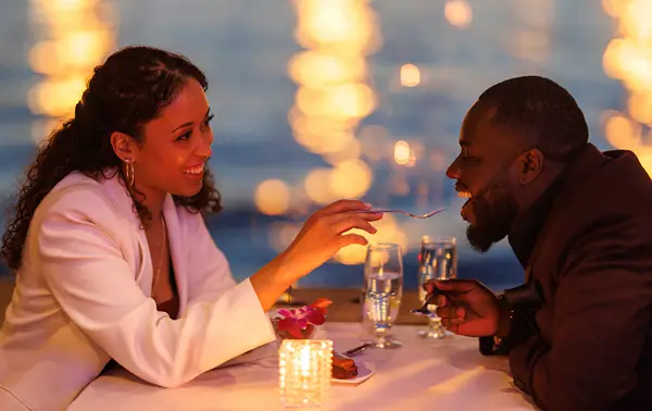 A couple sharing a romantic moment during a candlelit dinner by the water. The smiling woman feeds the man a bite of dessert, with blurred sparkling lights in the background.