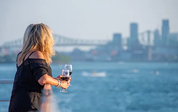 A blonde woman, viewed from behind, holding a glass of red wine, looking at a river with a bridge and skyscrapers in the background on a sunny day.