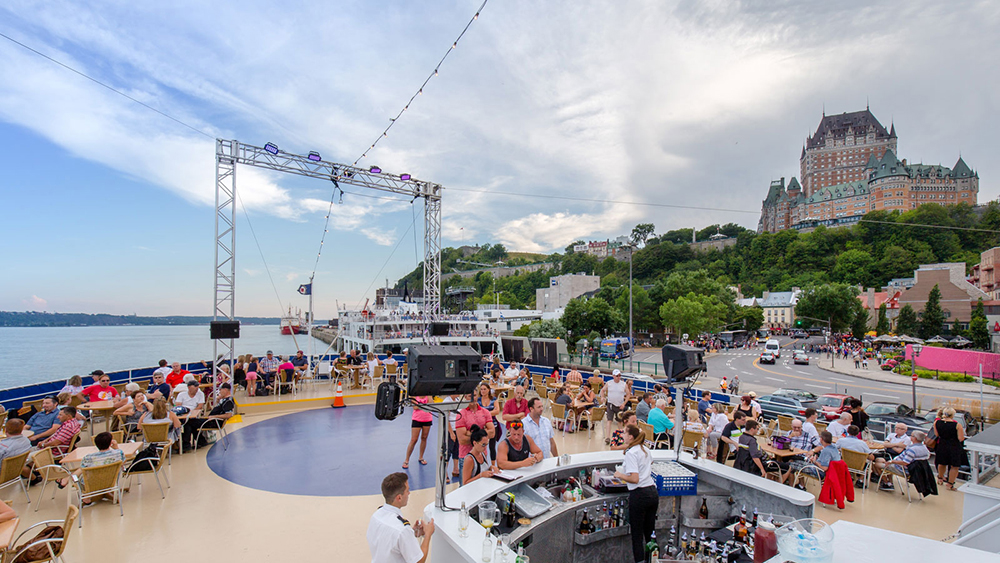 View of the front outdoor terrace of the AML Louis Jolliet boat, where customers are enjoying the view of the Saint Lawrence River, the Château Frontenac, and the panorama of Quebec City in the background