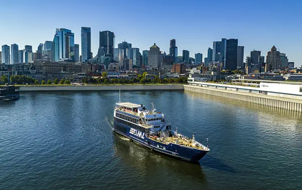A cruise ship sailing on a river, with a panoramic view of a city skyline featuring tall skyscrapers in the background under a clear sky.