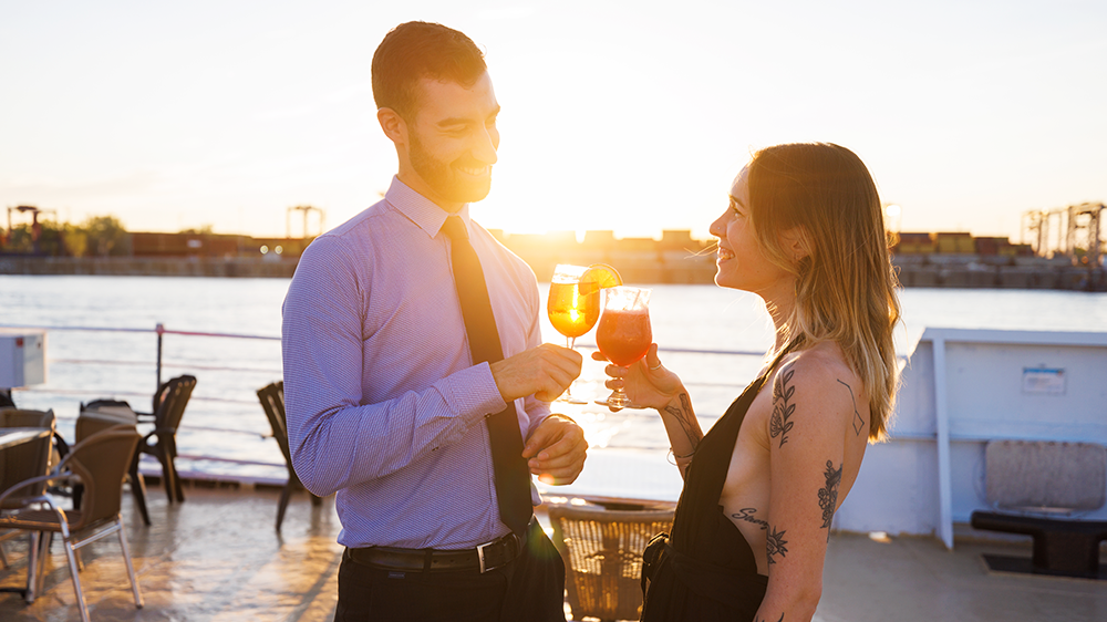 Couple complice et souriant buvant un cocktail sur la terrasse du navire AML Cavalier Maxim à Montréal avec un coucher de soleil en arrière plan