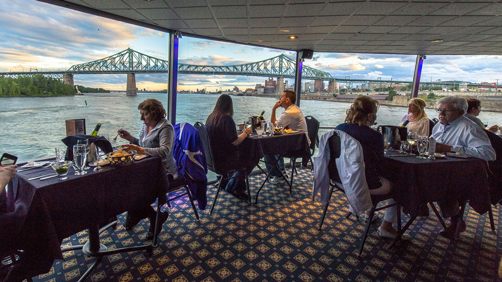 Guests are enjoying a bistronomic meal by the window aboard the AML Cavalier Maxim ship, with a stunning view of the Saint Lawrence River and the Jacques Cartier Bridge in Montreal in the background