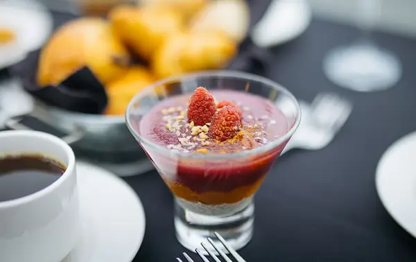 An elegant dessert in a glass, featuring layers of red fruit mousse or puree, topped with fresh raspberries and crumbled nuts or biscuits, accompanied by a cup of coffee and croissants in the background.