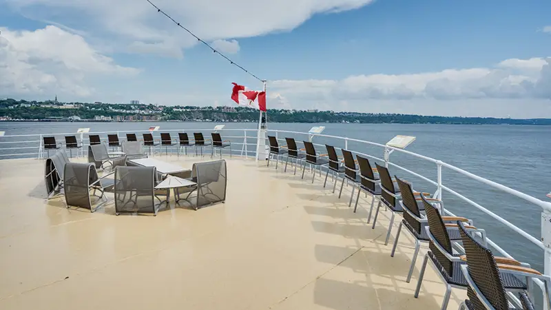 Front outdoor terrace of the AML Louis Jolliet ship in Quebec, with rows of chairs and tables, a Canadian flag waving, overlooking the Saint Lawrence River