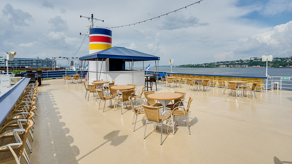 Upper deck outdoor terrace of the AML Louis Jolliet ship in Quebec, with tables and chairs available for customers, overlooking the Saint Lawrence River.