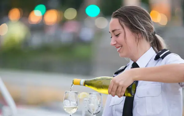A smiling waitress in uniform pours white wine into glasses, with a blurred background of lights and greenery.