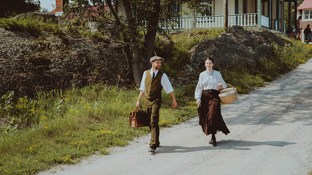 Two characters dressed in period costume are walking on Grosse Île. They are descending a small slope, with a historic house in the background.