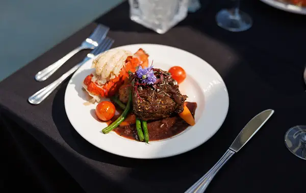 A gourmet plate featuring a filet of meat topped with sauce, served with green vegetables, cherry tomatoes, mashed potatoes, and an edible flower garnish, alongside grilled lobster. Presented on a black tablecloth with elegant cutlery.