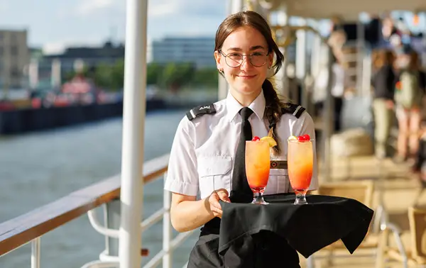 A waitress in uniform, wearing glasses and holding a tray with two colorful cocktails garnished with cherries. She stands on the deck of a boat, with a river and buildings in the background.