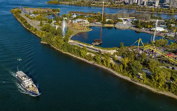 Aerial view of an island with an amusement park featuring roller coasters, a Ferris wheel, and rides, surrounded by a river. A cruise ship is sailing on the water to the left.