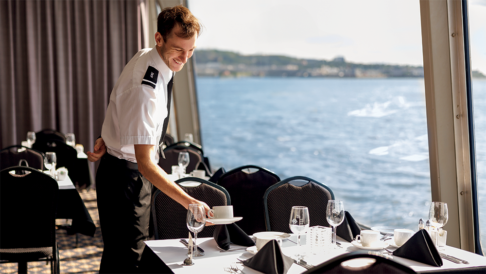 A smiling waiter in uniform setting a table near a large window overlooking the St. Lawrence River aboard the AML Cavalier Maxim in Montreal.