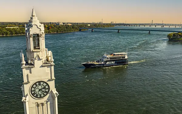 Aerial view of a tower with a large clock located by a river. A black cruise ship labeled "AML" sails on the water, with a bridge in the background and green riverbanks.