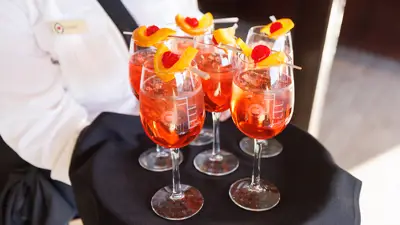 A server in uniform presents a tray of vibrant orange cocktails, garnished with orange slices and cherries on skewers, in elegant glasses.