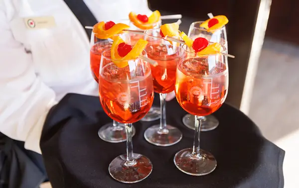 A server in uniform presents a tray of vibrant orange cocktails, garnished with orange slices and cherries on skewers, in elegant glasses.