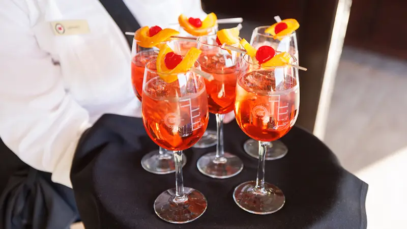 A server in uniform presents a tray of vibrant orange cocktails, garnished with orange slices and cherries on skewers, in elegant glasses.