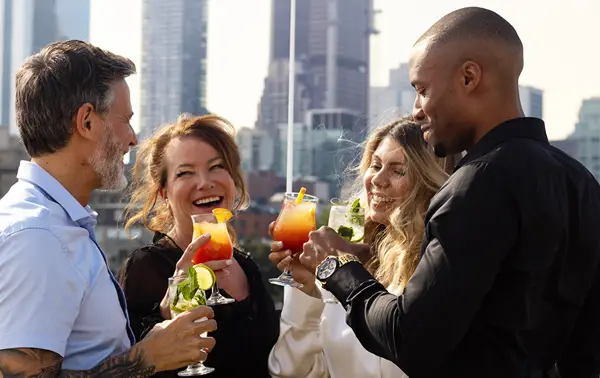 A group of four smiling friends toasting with colorful cocktails on a terrace, with skyscrapers in the background.