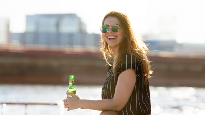 A smiling woman wearing sunglasses and a striped shirt is holding a bottle of beer on the terrace of the AML Cavalier Maxim ship in Montreal.