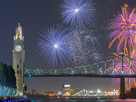 Colorful fireworks burst in the night sky near the Jacques Cartier Bridge and the Clock Tower, with reflections on the river and a crowd watching from the quay.