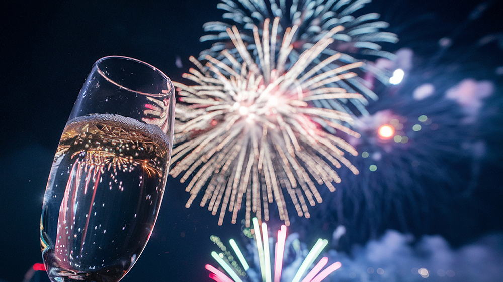 A champagne glass raised in the foreground while multicolored fireworks light up the night sky in the background.