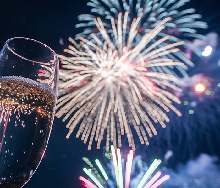 A champagne glass raised in the foreground while multicolored fireworks light up the night sky in the background.