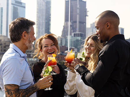 A group of four smiling friends toasting with colorful cocktails on a terrace, with skyscrapers in the background.