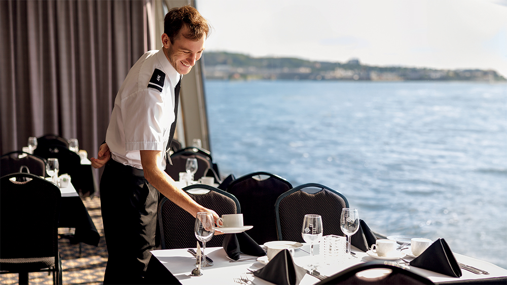 A uniformed waiter sets an elegant table with white tablecloths and black napkins in a dining room with a panoramic view of the water in the background.