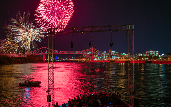 A vibrant fireworks display in a black sky above the AML Cavalier Maxim ship in Montreal, with the city illuminated in the distance.