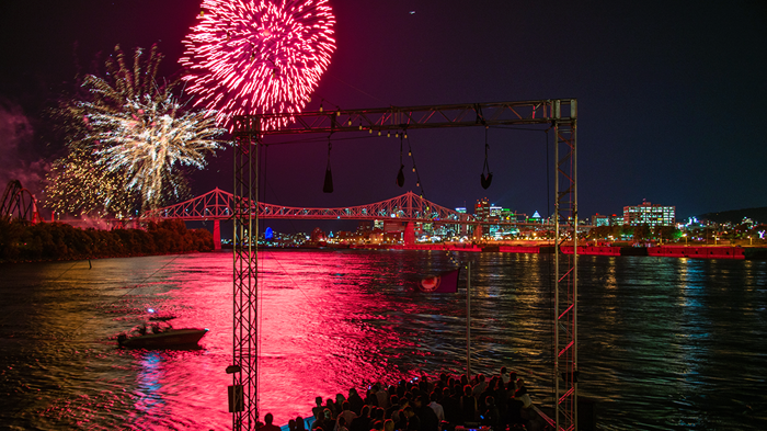 A vibrant fireworks display in a black sky above the AML Cavalier Maxim ship in Montreal, with the city illuminated in the distance.