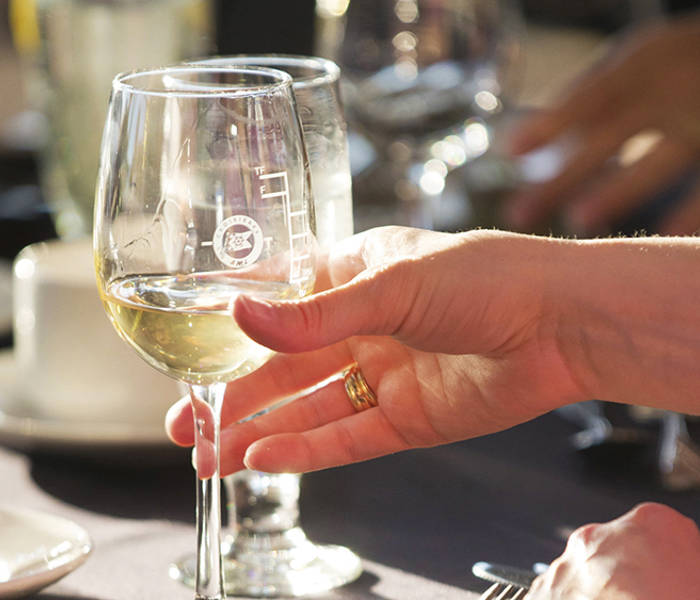 A hand holds a glass of white wine illuminated by sunlight, with an elegant table in the background featuring cups and glasses.