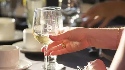 A hand holds a glass of white wine illuminated by sunlight, with an elegant table in the background featuring cups and glasses.