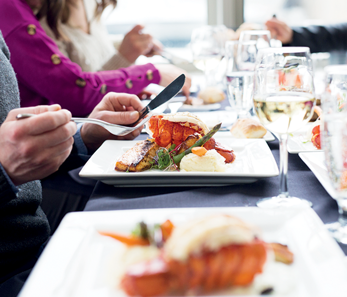 Gros plan sur des gens qui dînent avec une assiette de homard au premier plan accompagnée de verres de vin et d'autres plats autour. 