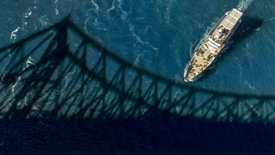 Aerial view of a boat sailing under a bridge, with the bridge's shadow cast on the blue surface of the water.