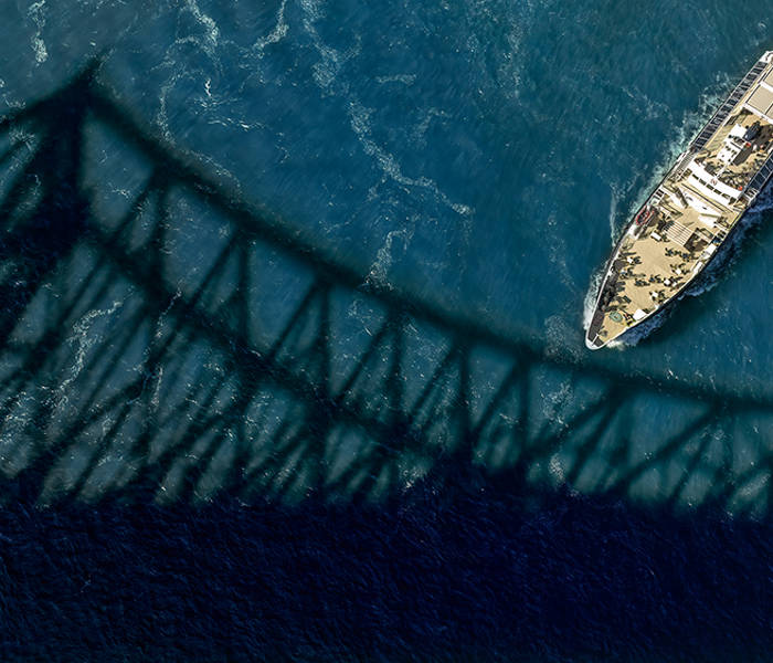 Aerial view of a boat sailing under a bridge, with the bridge's shadow cast on the blue surface of the water.