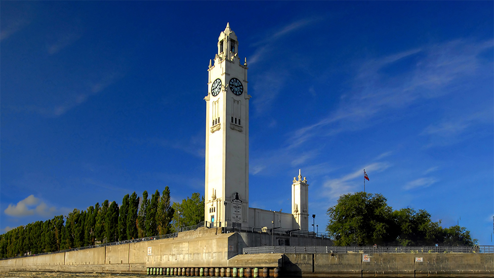 Une grande tour horloge blanche située sur un quai, entourée d'arbres et sous un ciel bleu dégagé. Une petite structure et un drapeau sont visibles à proximité.