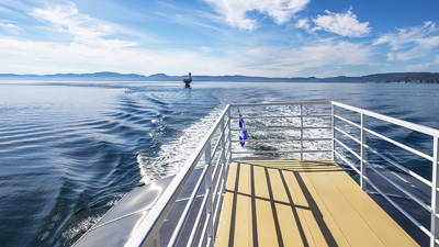 View from the back of a boat, showing a sunlit deck with metal railings. The boat's wake ripples across the calm water, and a lighthouse is visible in the distance. Hills on the horizon under a blue sky with light clouds complete the scenery.