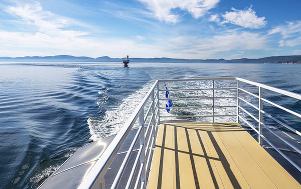 View from the back of a boat, showing a sunlit deck with metal railings. The boat's wake ripples across the calm water, and a lighthouse is visible in the distance. Hills on the horizon under a blue sky with light clouds complete the scenery.
